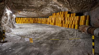 Das Bild zeigt einen unterirdischen Lagerraum, in dem zahlreiche gelbe Fässer gestapelt sind. Der Raum scheint in einem Felsgestein ausgehoben zu sein und wird durch künstliches Licht beleuchtet. Im Vordergrund ist ein Bereich durch eine Kette und Warnschilder abgesperrt, die auf radioaktive Materialien hinweisen. Die Fässer sind ordentlich gestapelt und erstrecken sich entlang der Wände des Lagerraums.