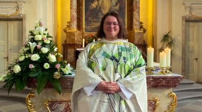 Pfarrerin Sabine Clasani steht vor dem Altar. Sie trägt ein weiß-grünes Gewand und lächelt.