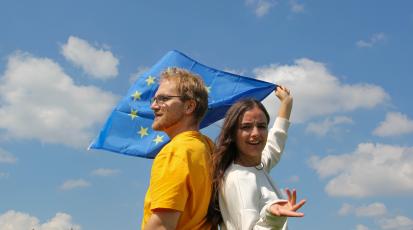 Ein Mann und eine Frau stehen Rücken an Rücken und schauen kritisch. Sie halten eine EU-Flagge hinter sich