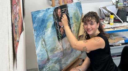 Die Kunststudentin Ines sitzt in dem Atelier ihrer Hochschule und malt mit einem Pinsel ein Kunstwerk auf Leinwand. Dabei schaut sie lächelnd in die Kamera.