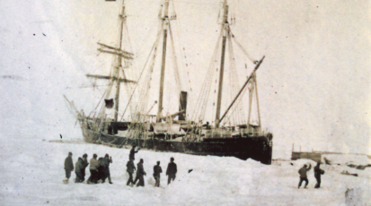 Zu sehen ist ein großes, altes Expeditionsschiff, das im Eis gefangen scheint. Davor stehen Menschen auf dem Eis.