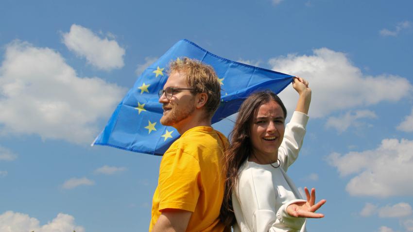 Ein Mann und eine Frau stehen Rücken an Rücken und schauen kritisch. Sie halten eine EU-Flagge hinter sich.