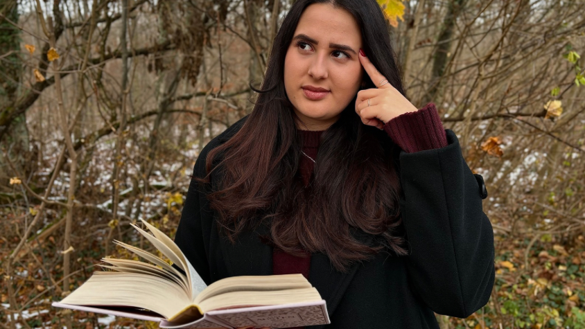 Eine Person liest ein Buch in der Natur und hat einen fragenden Gesichtsausdruck. 