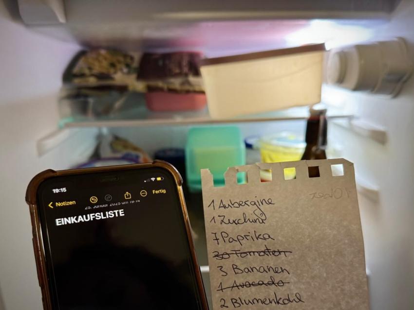 Auf einem Smartphone wird eine Einkaufsliste erstellt. Daneben ist eine handschriftliche Einkaufsliste auf einem Notizzettel. Im Hintergrund erkennt man Lebensmittel in einem Kühlschrank.