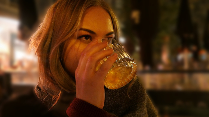 Eine Frau, die draußen in einer Bar sitzt und ein Glas mit Whiskey zum trinken ansetzt.