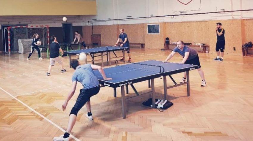 In einer Sporthalle stehen zwei Tischtennisplatten. Eine steht im Vordergrund, an dieser sind zwei Spieler in normalen Sportklamotten zu sehen. Sie lehnen sich über den Tisch und versuchen, einen Volleyball mit dem Kopf zu spielen. Der Ball wird von dem linken Spieler gerade mit dem Kopf gespielt.