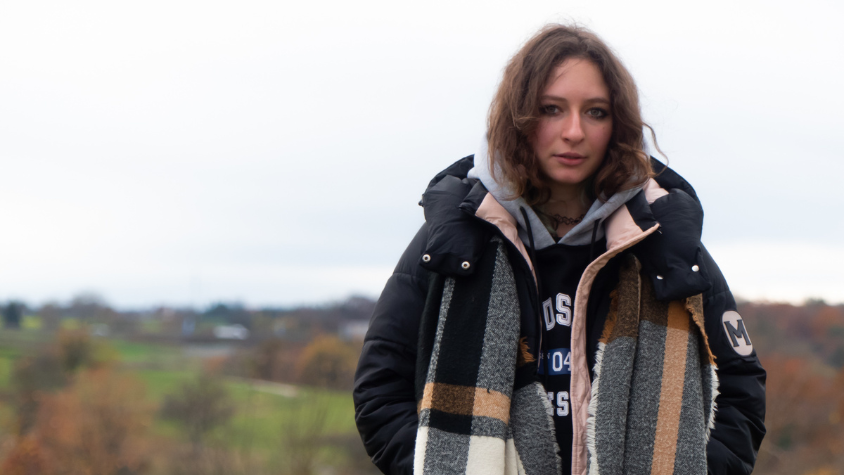 Junge Frau, die starr in die Kamera bilckt. Im Hintergrund befinden sich grüne Felder und leicht düsterer Himmel.