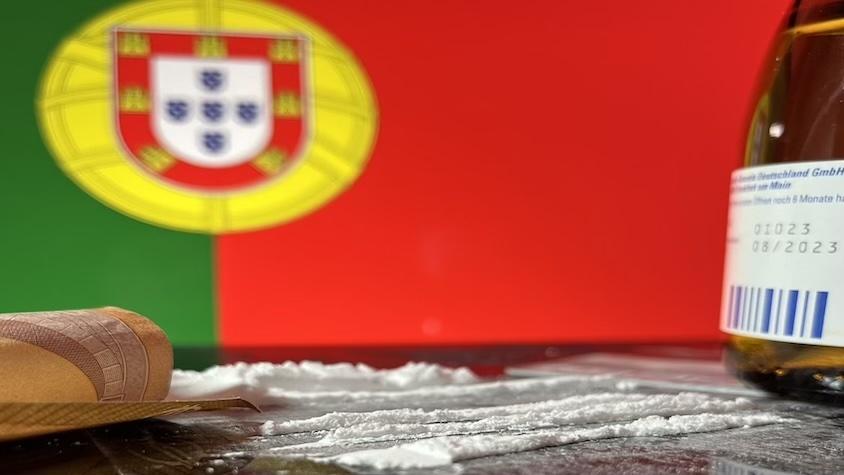 Gerollter 50-Euro-Schein liegt vor Kokainbahnen, um so Drogenkonsum anzudeuten. Die Flagge Portugals ist im Hintergrund zu sehen und an der rechten Seite ist eine Flasche von codeinhaltigem Hustensaft zu sehen.