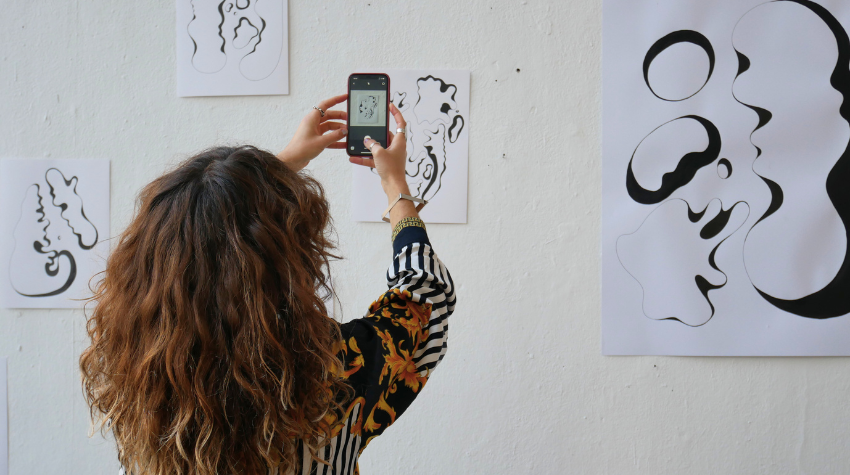 Junge Frau fotografiert mit ihrem Smartphone ein Kunstwerk, das an der Wand hängt.