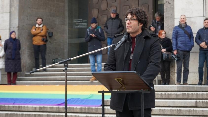 Alon Bindes, ein Mann mit dunklen Locken, Brille und schwarzem Mantel hält eine Rede. 