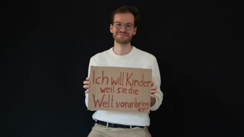 Es ist ein junger Mann mit einem Schild zu sehen. Auf dem Schild steht: Ich will Kinder, weil sie die Welt voranbringen.  